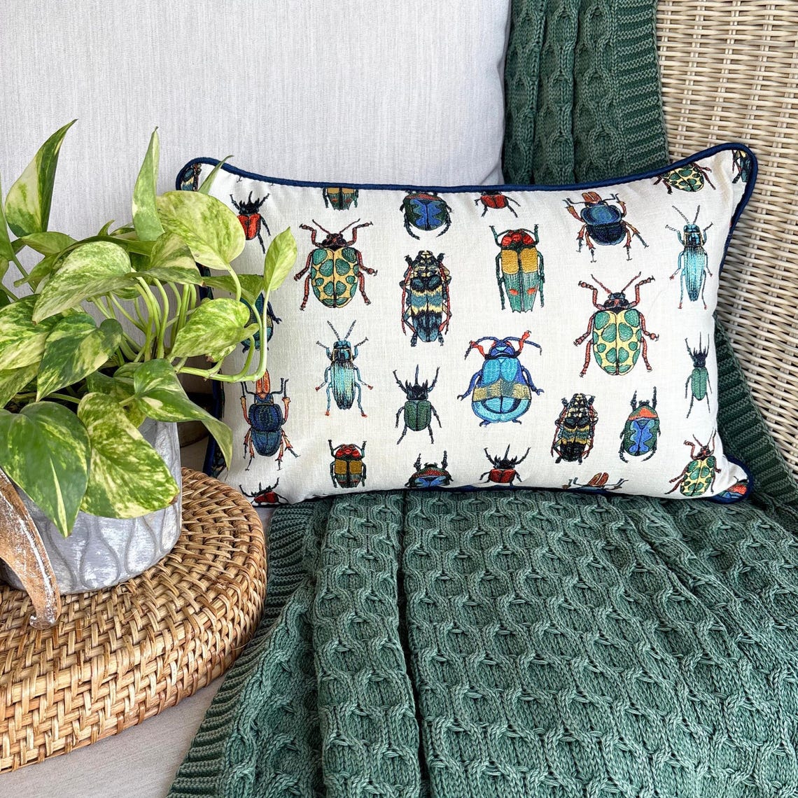 A decorative lumbar cushion cover with a blue background featuring a pattern of various colorful insects, displayed on a chair with a skeleton and Halloween decorations around it.